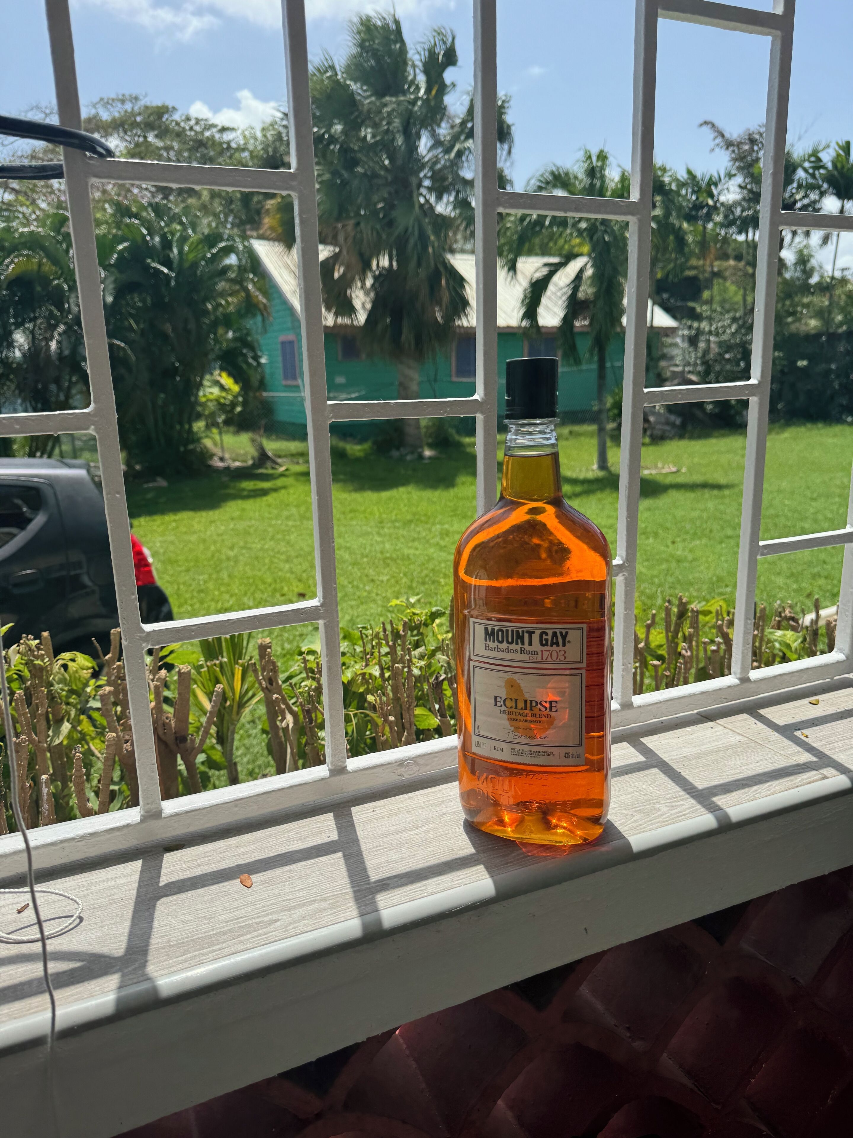 A bottle of Mount Gay Barbados Rum on a windowsill with a view of a green lawn and palm trees.