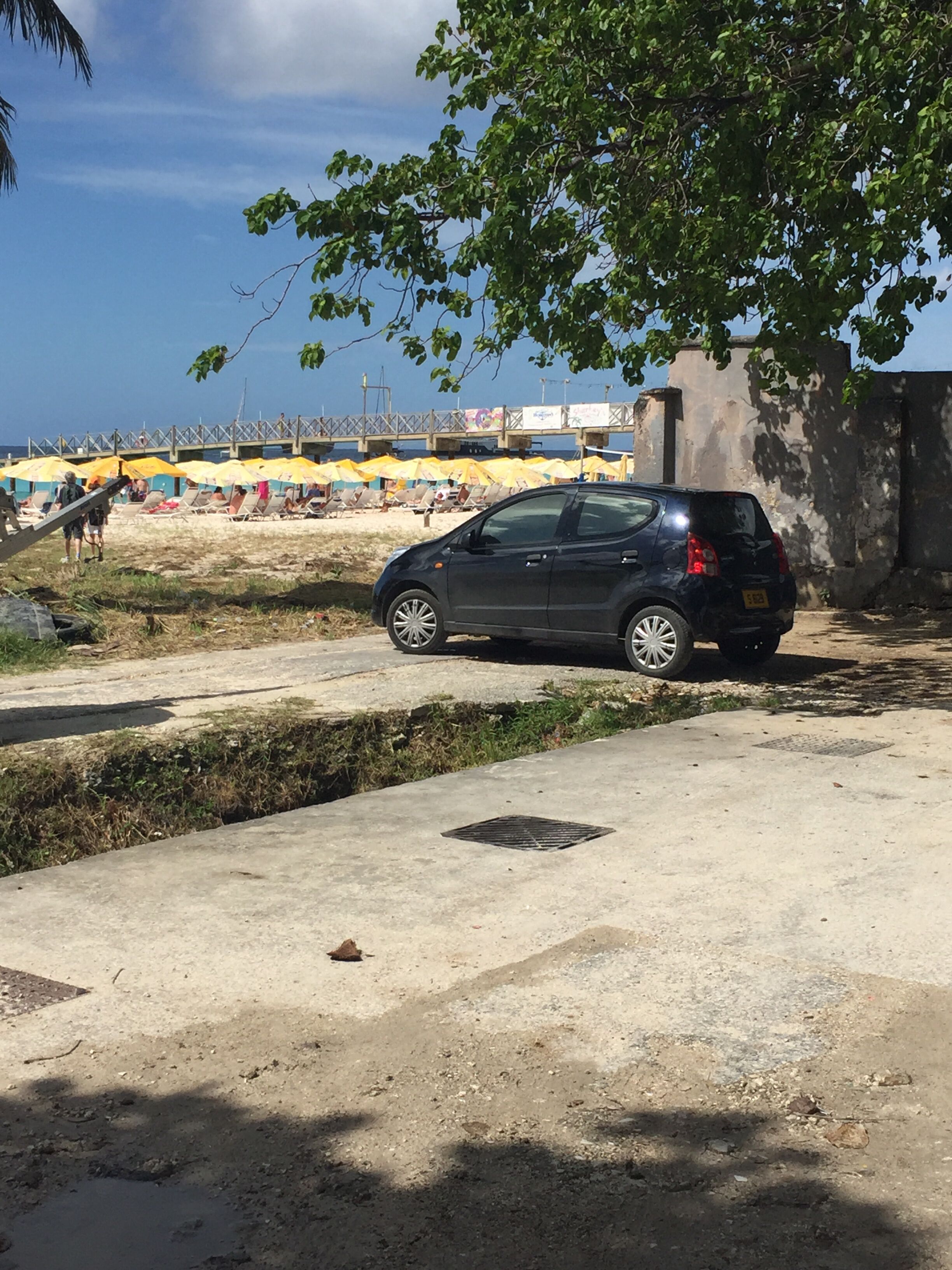 A black car parked on a concrete surface near a beach with yellow umbrellas.