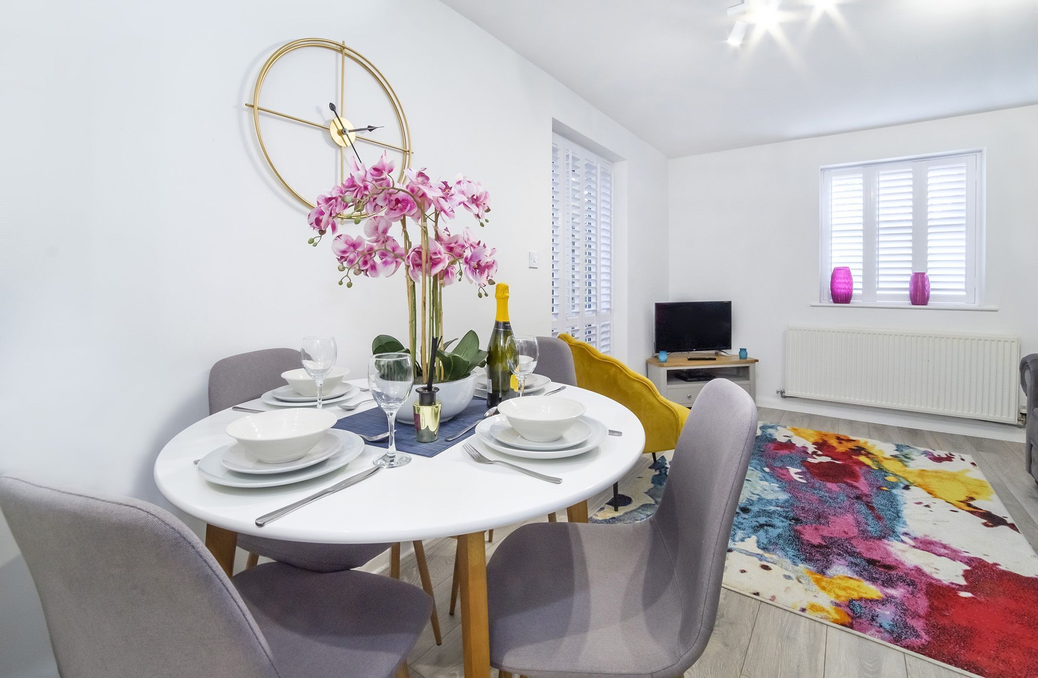 A dining area with a round table set for four, a gold clock on the wall, and a colorful rug on the floor.