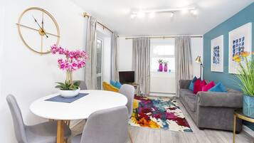 A modern living room with a round dining table, grey sofas, a colorful rug, and a large window with curtains.