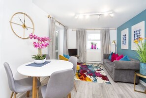 Un salon moderne avec une table à manger ronde, des canapés gris, un tapis coloré et une grande fenêtre avec des rideaux.
