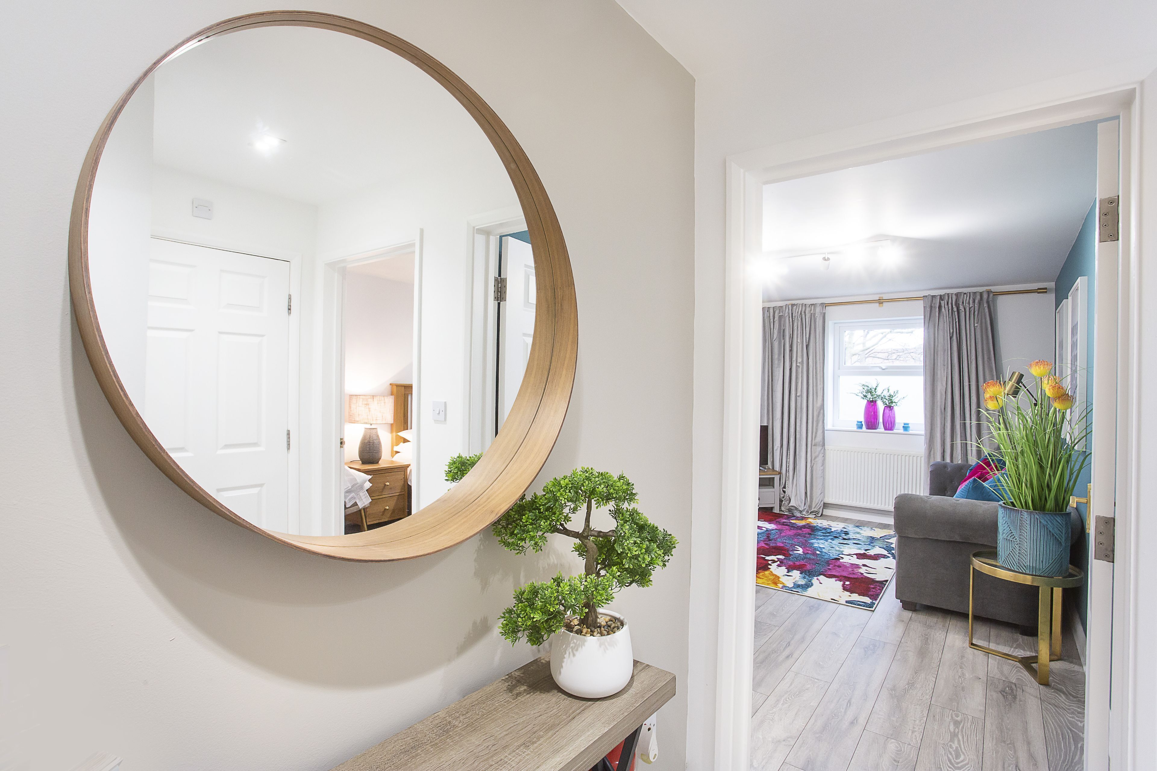 A hallway with a round mirror, a potted plant, a wooden console table, and a glimpse of a living room with a sofa and a window.