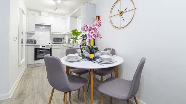 A modern kitchen with white cabinets, a microwave, and an oven. There is a dining table with chairs and a vase of flowers. - Minster's Reach Holiday Apartment (YORK)
