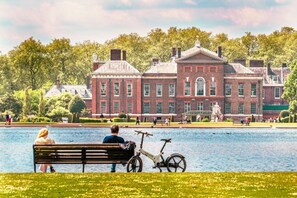 Un parque con un lago, una banca, una bicicleta y un edificio de ladrillo grande con arquitectura clásica.