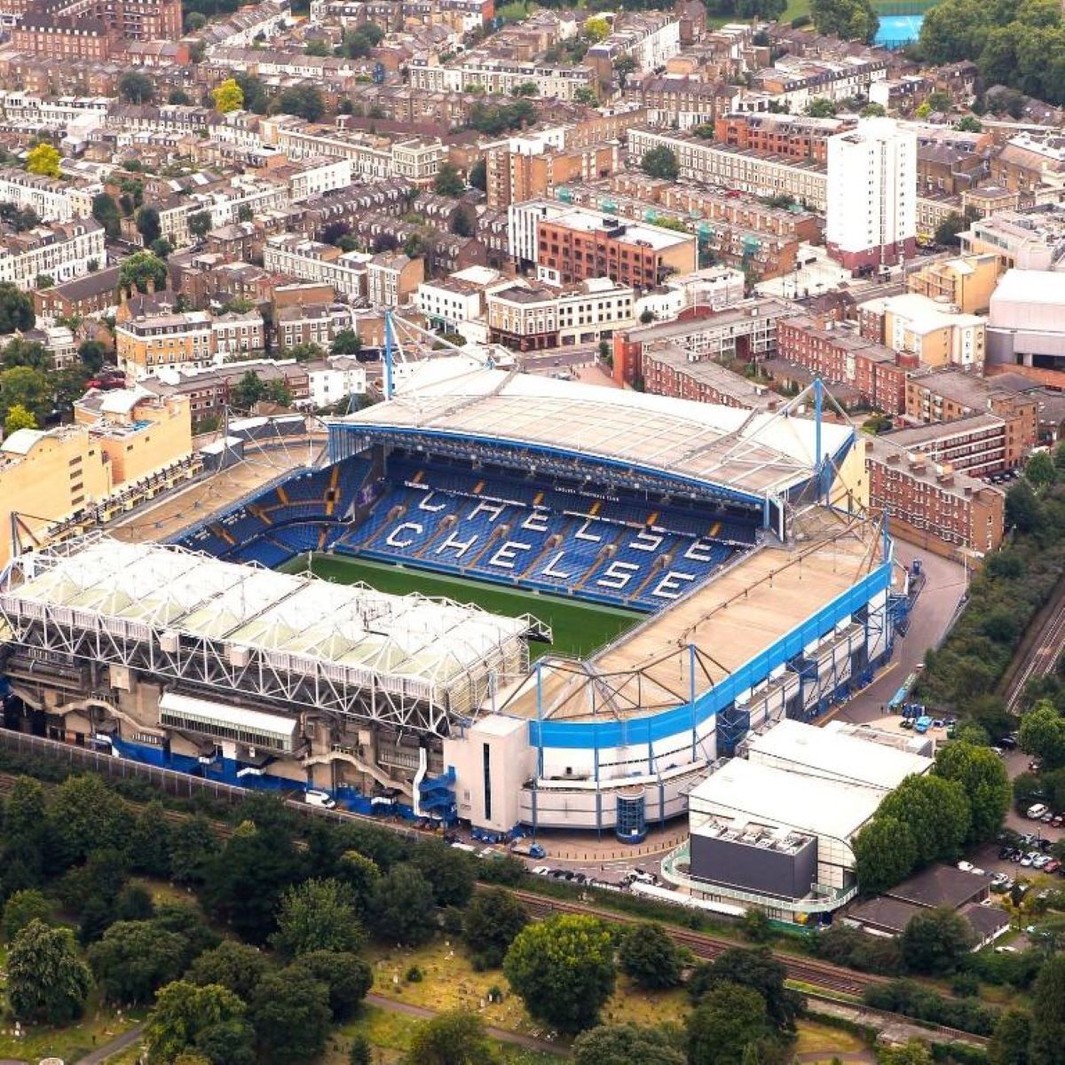 An aerial view of a stadium with "CHELSEA" written on the field, surrounded by urban buildings.