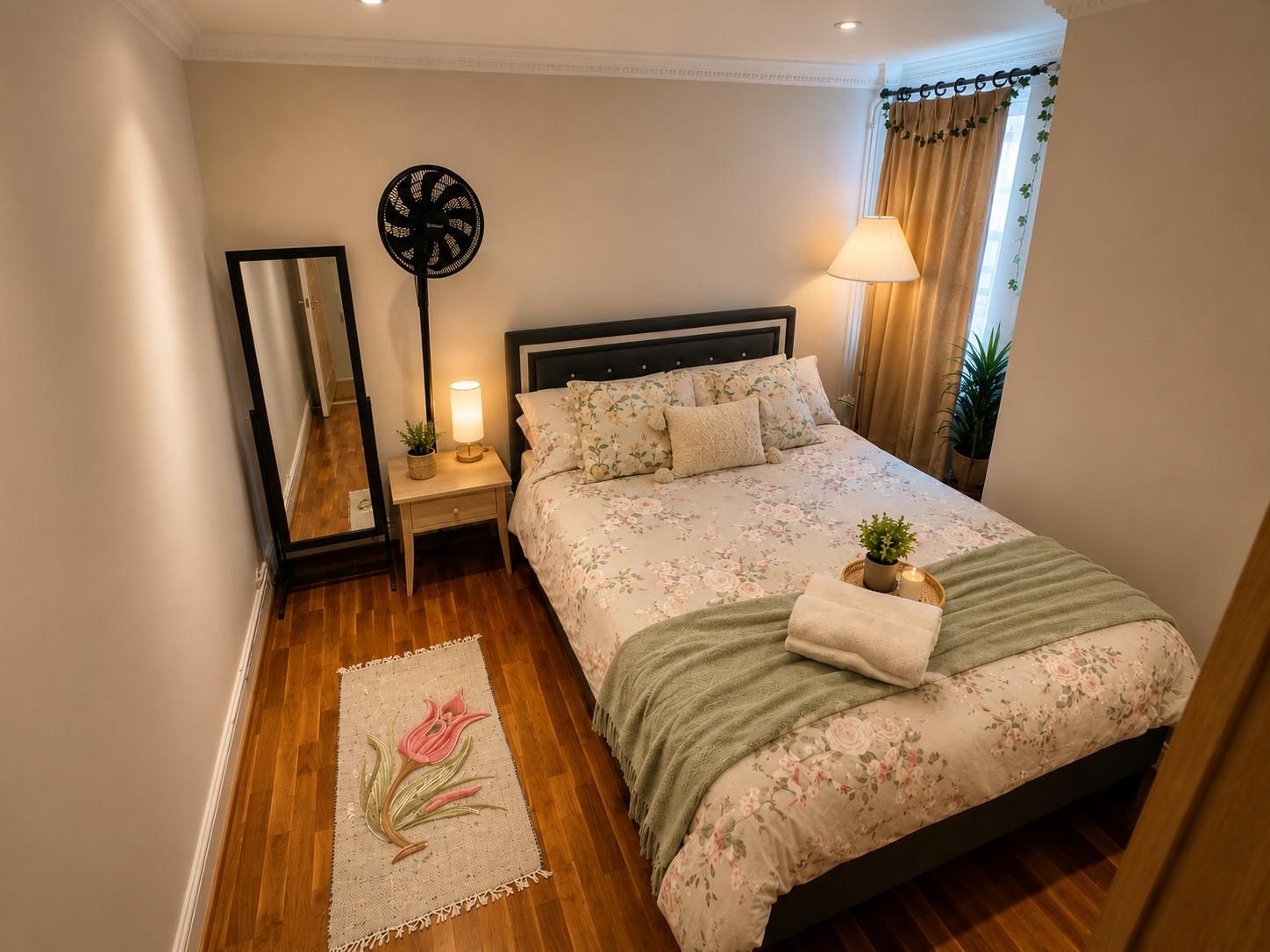 Een slaapkamer met een bed, nachtkastje, lamp, spiegel en een vloerkleed met bloemen.