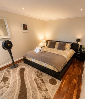 A bedroom with a bed, a standing fan, a nightstand, and a wall-mounted lamp.