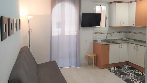 Una cocina compacta con gabinetes blancos, un taburete pequeño y un televisor de pantalla plana sobre el lavamanos.