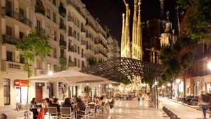 Escena urbana nocturna con una zona de café que ofrece mesas al aire libre bajo una cubierta, una catedral destacada con agujas doradas y una fila de edificios de varios pisos con balcones.