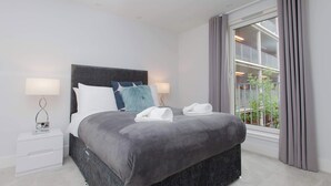 A modern hotel room with a large bed, bedside lamps, a nightstand, and a view of greenery through the window. - Luxurious Central Edinburgh Apartment - Sleeps 6 (Edinburgh)