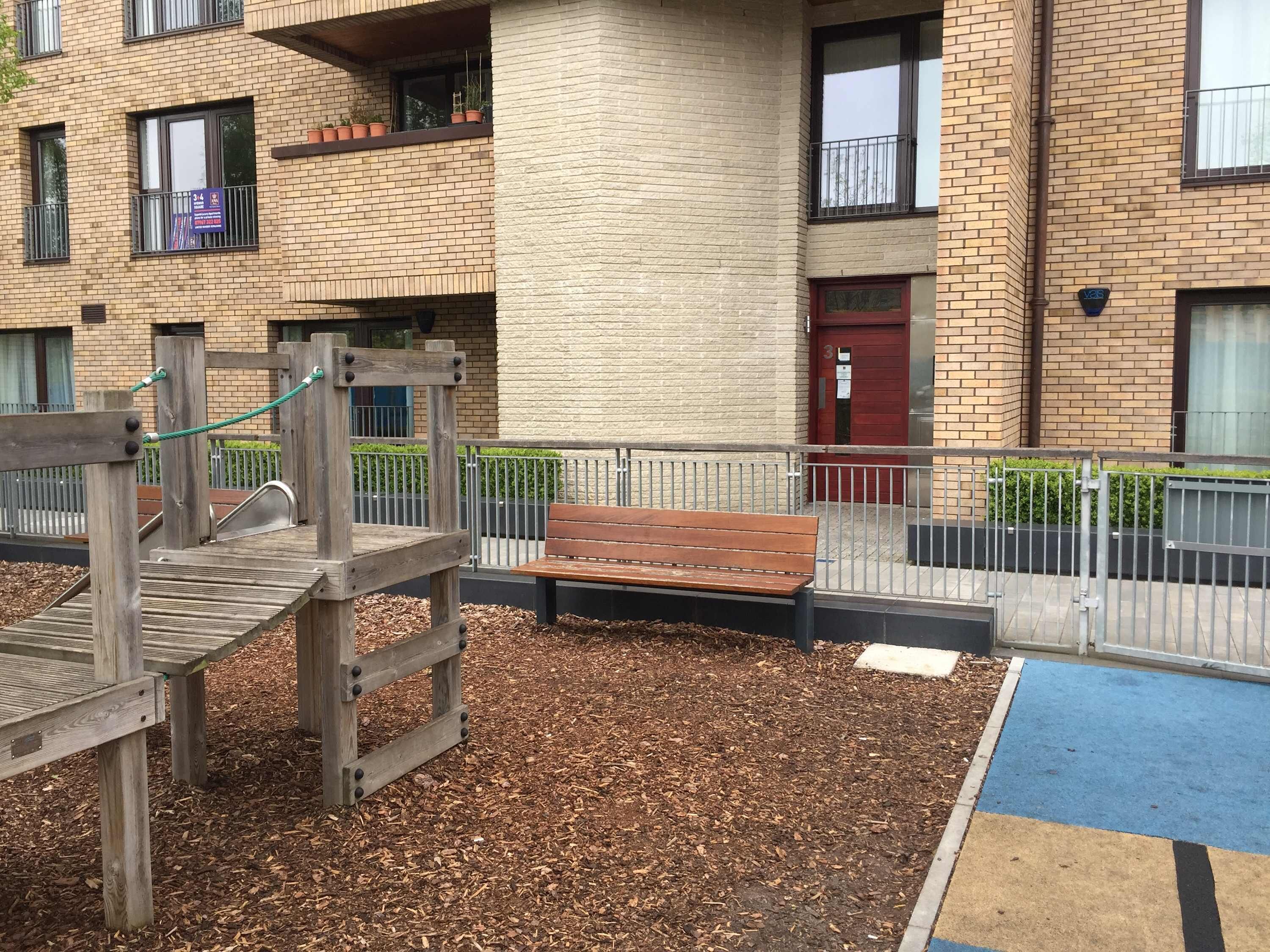 Een speeltuin met houten speeltoestellen en een bankje, omringd door een bakstenen gebouw met ramen en een rode deur.