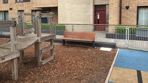A playground with wooden play structures and a bench, surrounded by a brick building with windows and a red door. - Luxurious Central Edinburgh Apartment - Sleeps 6 (Edinburgh)