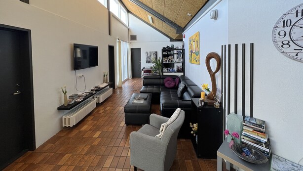 A spacious living room with a large wall clock, a black leather sofa, a grey armchair, and a bookshelf filled with books and decorative items. - Godaland Guesthouse (Rangárþing eystra)