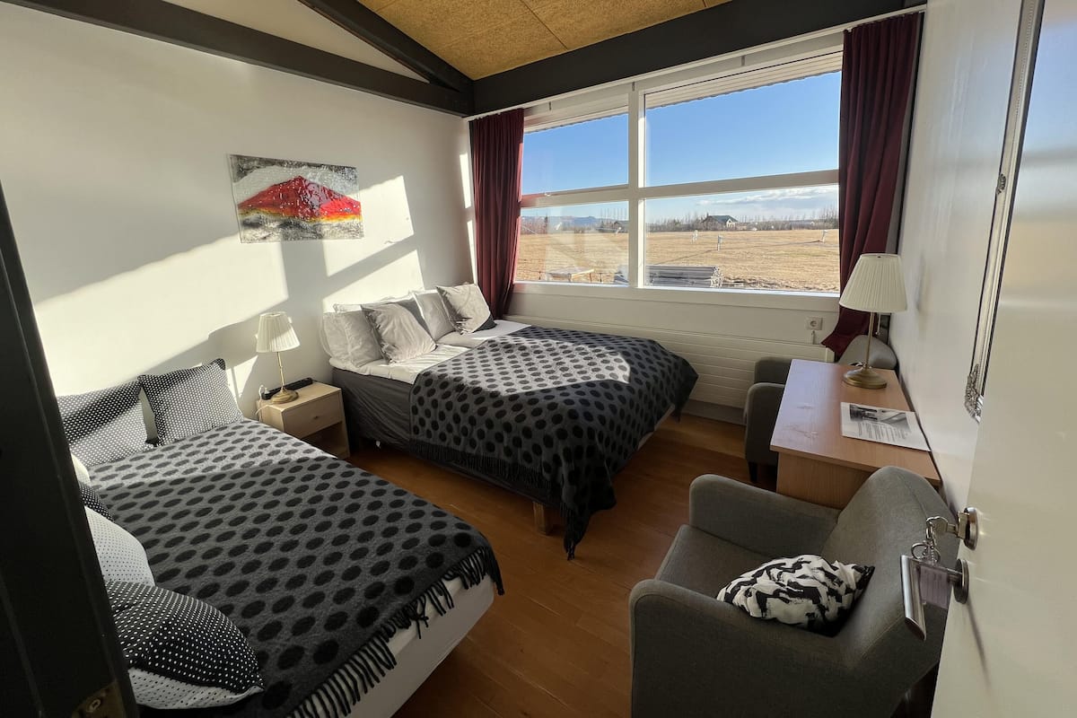 A room with two beds, a desk, and a chair. There is a window with curtains, a lamp, and a painting on the wall.