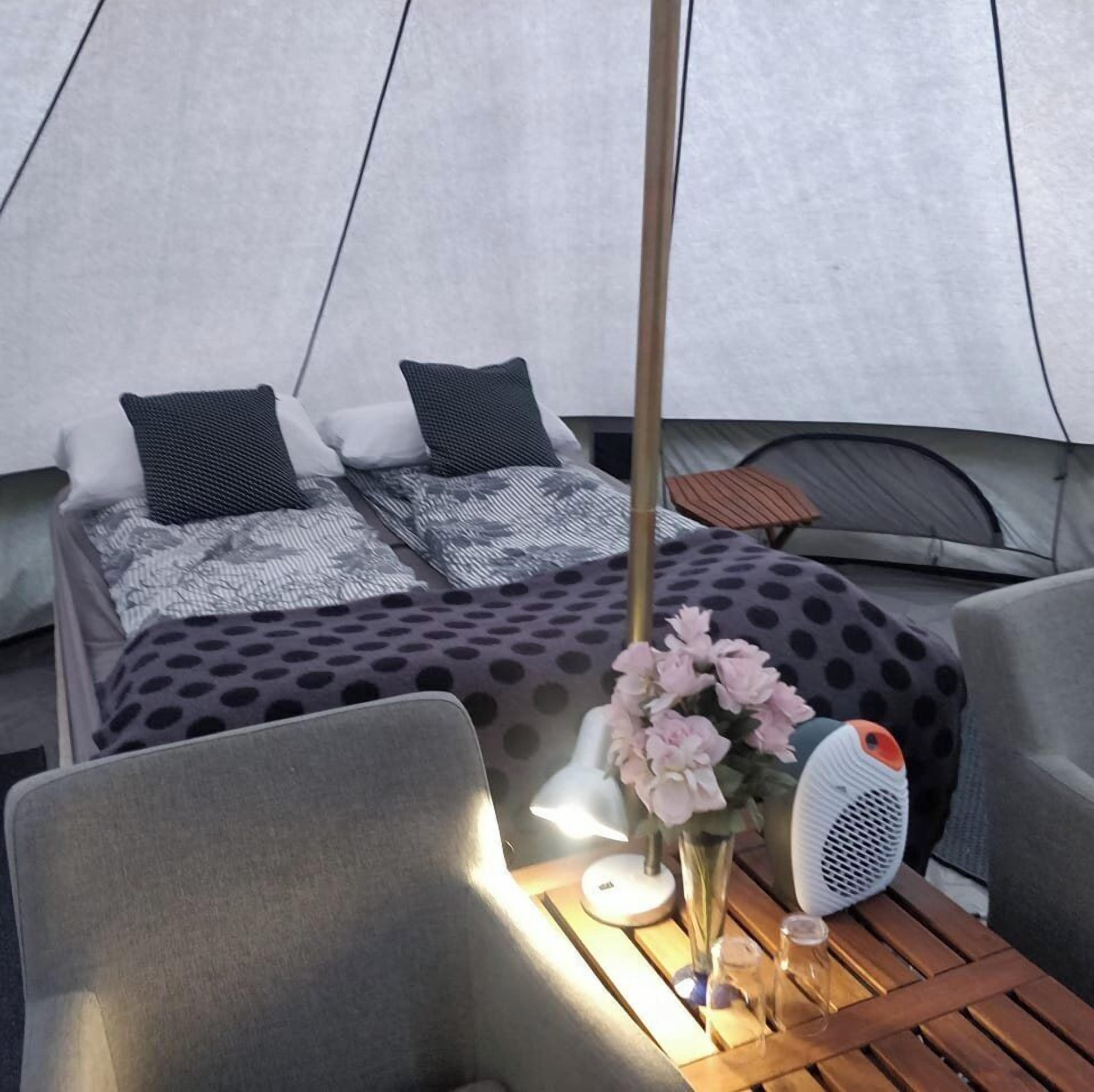 A tented accommodation with a bed, pillows, a polka dot blanket, a vase with flowers, a fan, and a table with glasses.