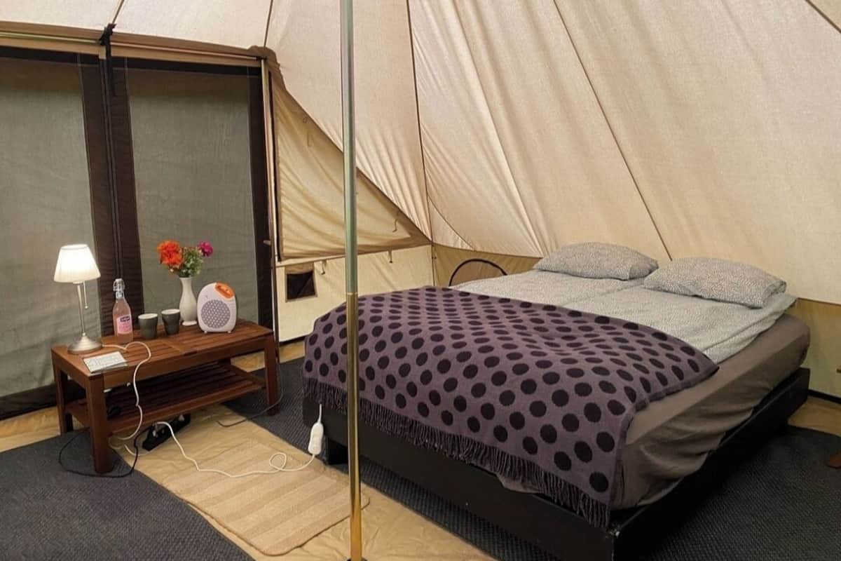 A tent with a bed, a nightstand with a lamp, a vase with flowers, and a speaker.