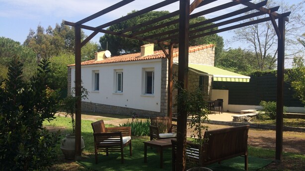 Ein überdachter Außenbereich mit einer Pergola, ein Steinhaus mit Ziegeldach und eine grüne Rasenfläche.