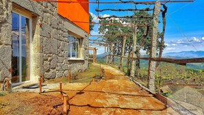 Un bâtiment en pierre doté d’une fenêtre et d’une porte, une pergola en bois recouverte de plantes grimpantes, et une vue sur la campagne.
