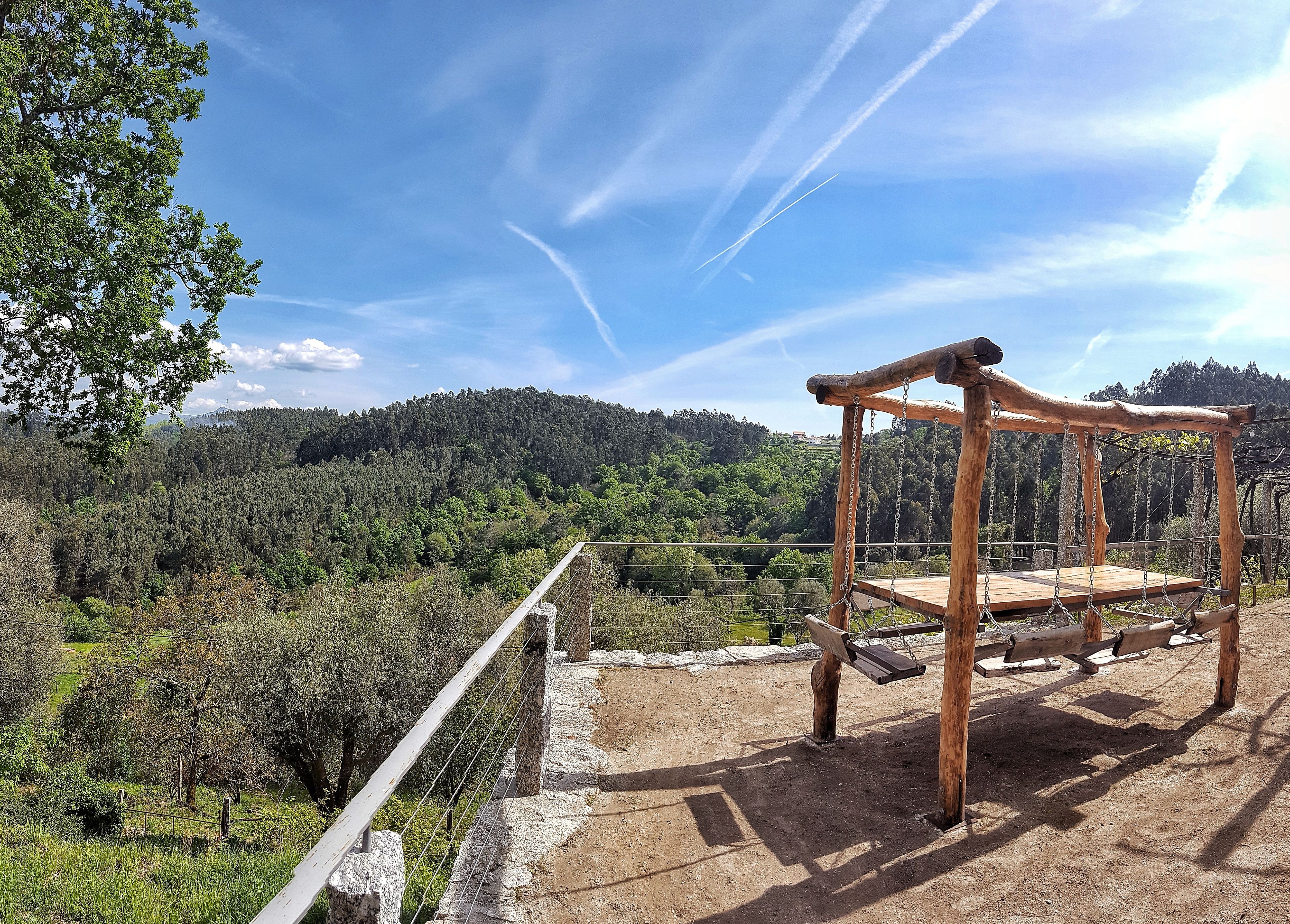 Une balançoire en bois rustique, installée sur un chemin de terre, offre une vue sur une colline boisée et un ciel dégagé.
