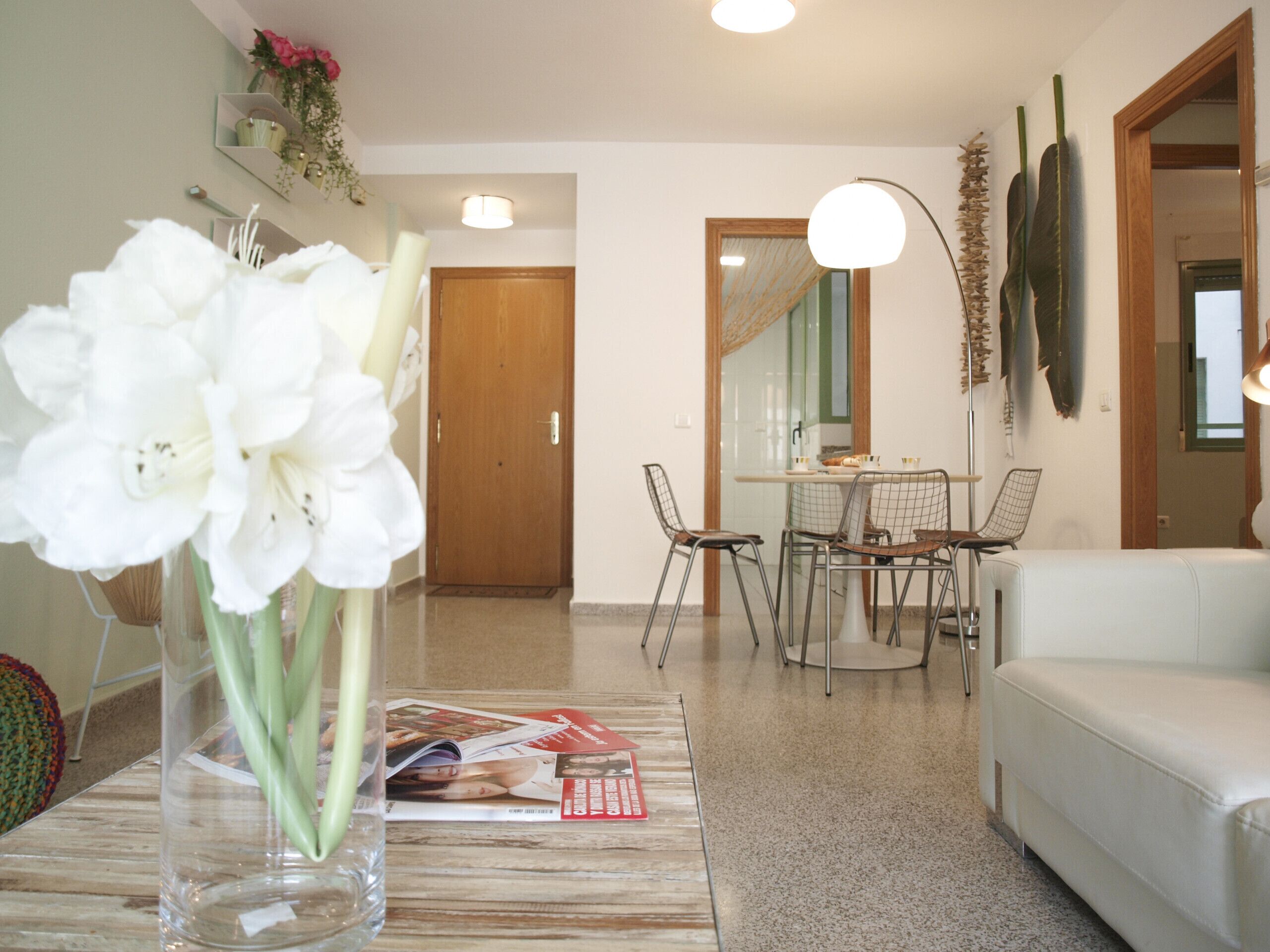 Ein Wohnzimmer mit einem weißen Sofa, einem Esstisch mit Stühlen, einer Vase mit weißen Blumen und einer Zeitschrift auf dem Tisch.