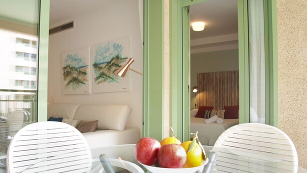Un balcone con un tavolo su cui sono disposti della frutta, sedie bianche e vista su una camera da letto con un letto e tende.
