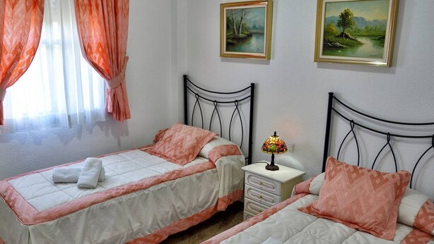 A hotel room with two beds, each with a white and orange bedspread, a bedside table with a lamp, and two framed landscape paintings on the wall. - Chalet el Naranjo - Sevilla (Dos Hermanas)