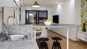 Eine moderne Küche mit Granit Arbeitsplatten, einer Edelstahlspüle und einer Zitronenschale auf der Arbeitsfläche. Zwei schwarze Hocker und ein weißer Stuhl sind vorhanden. Eine grüne Pendelleuchte hängt über der Arbeitsfläche.