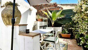 Une petite terrasse extérieure avec une table et des chaises, entourée de plantes en pot et agrémentée d’un grand parasol qui offre de l’ombre.