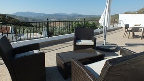 Terraço com móveis de vime, uma mesa e vista para montanhas distantes.