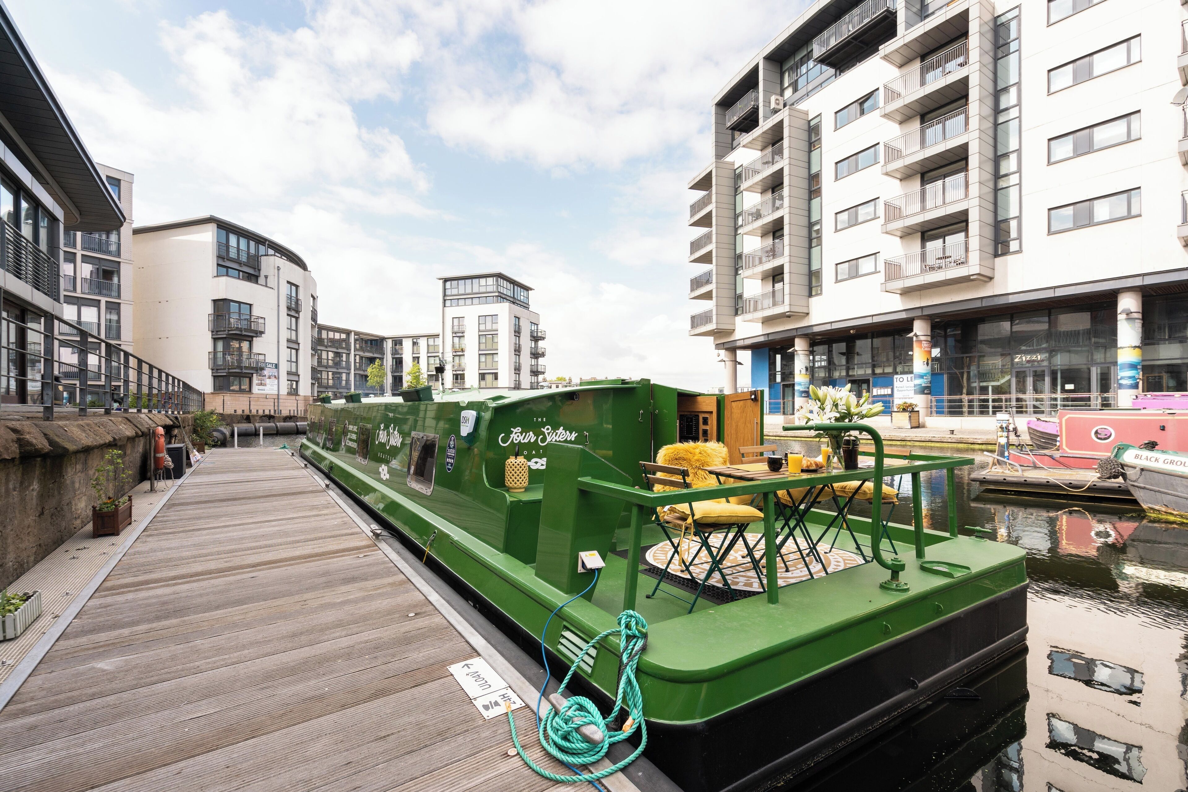 Un barco verde atracado en un muelle con una pasarela de madera, rodeado de edificios modernos y un cielo despejado.