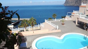 Ein Poolbereich mit Blick auf das Meer, Palmen und ein Gebäude mit einem weißen Balkon.