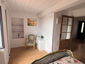 Une pièce comprenant un lit, une chaise, une petite table, un tableau et une porte donnant sur une autre pièce.