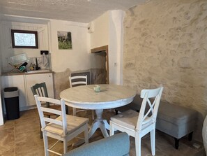 Un coin repas comprenant une table ronde et quatre chaises, un banc, une poubelle et un petit espace cuisine avec une fenêtre et un tableau au mur.