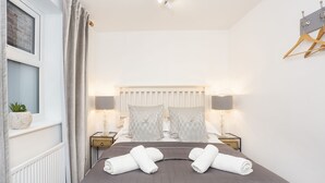Un lit bien fait, recouvert d’une couverture grise, deux tables de chevet avec des lampes, une fenêtre avec des rideaux et un porte-manteau fixé au mur.