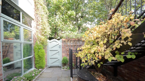 Un bâtiment en briques avec une porte en verre, un escalier doté d’une rampe en métal et un espace jardin agrémenté de fleurs et d’un arbre.