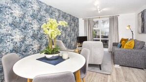 Un salon moderne avec une table à manger ronde, des chaises recouvertes d’un tissu gris, un canapé gris agrémenté de coussins jaunes, un téléviseur et une grande fenêtre avec des rideaux.