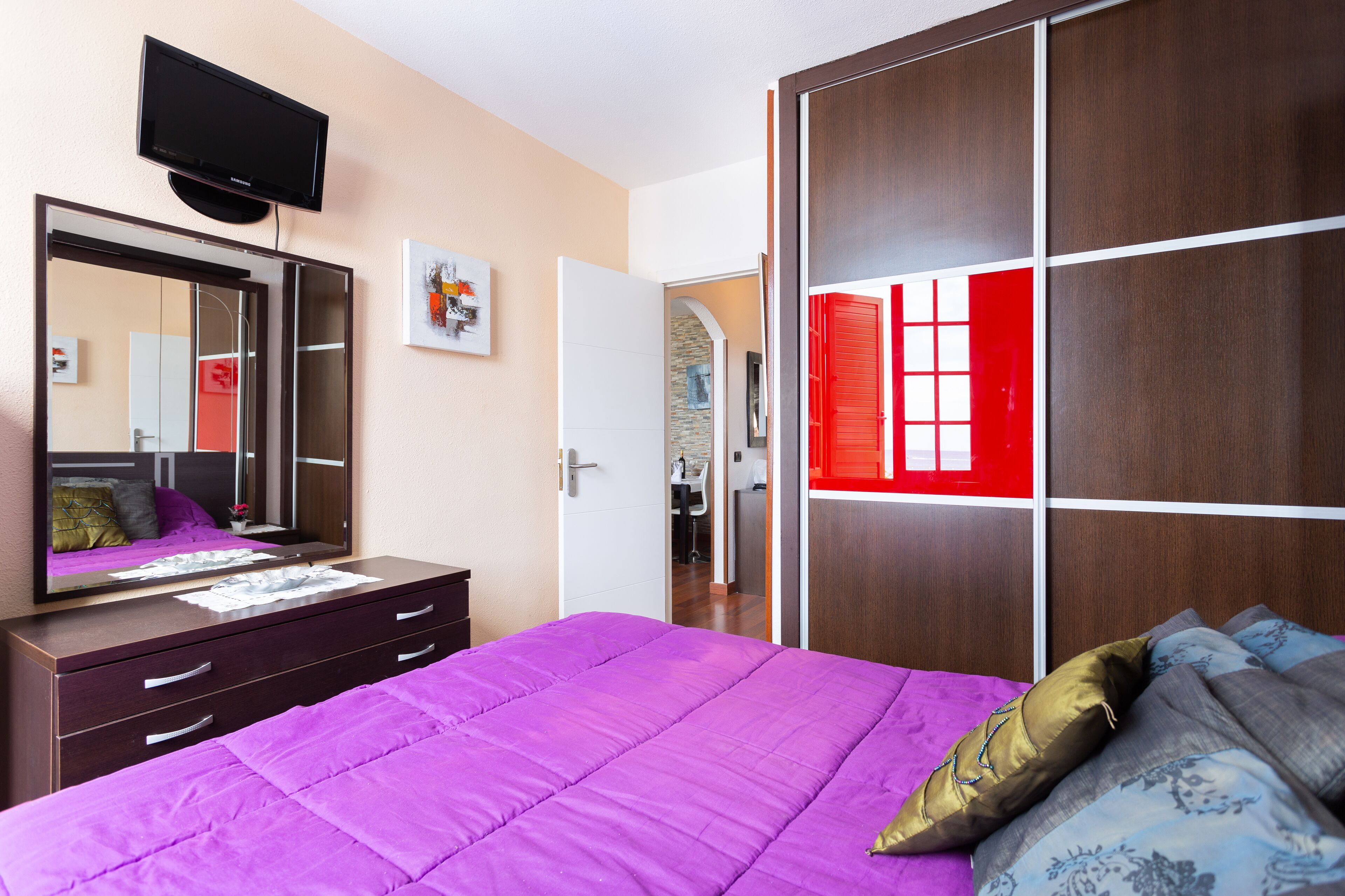 Une chambre avec une couvre-lit violette, un grand miroir, un téléviseur fixé au-dessus de la commode et une armoire aux panneaux rouges et blancs.