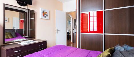 Une chambre avec une couvre-lit violette, un grand miroir, un téléviseur fixé au-dessus de la commode et une armoire aux panneaux rouges et blancs.