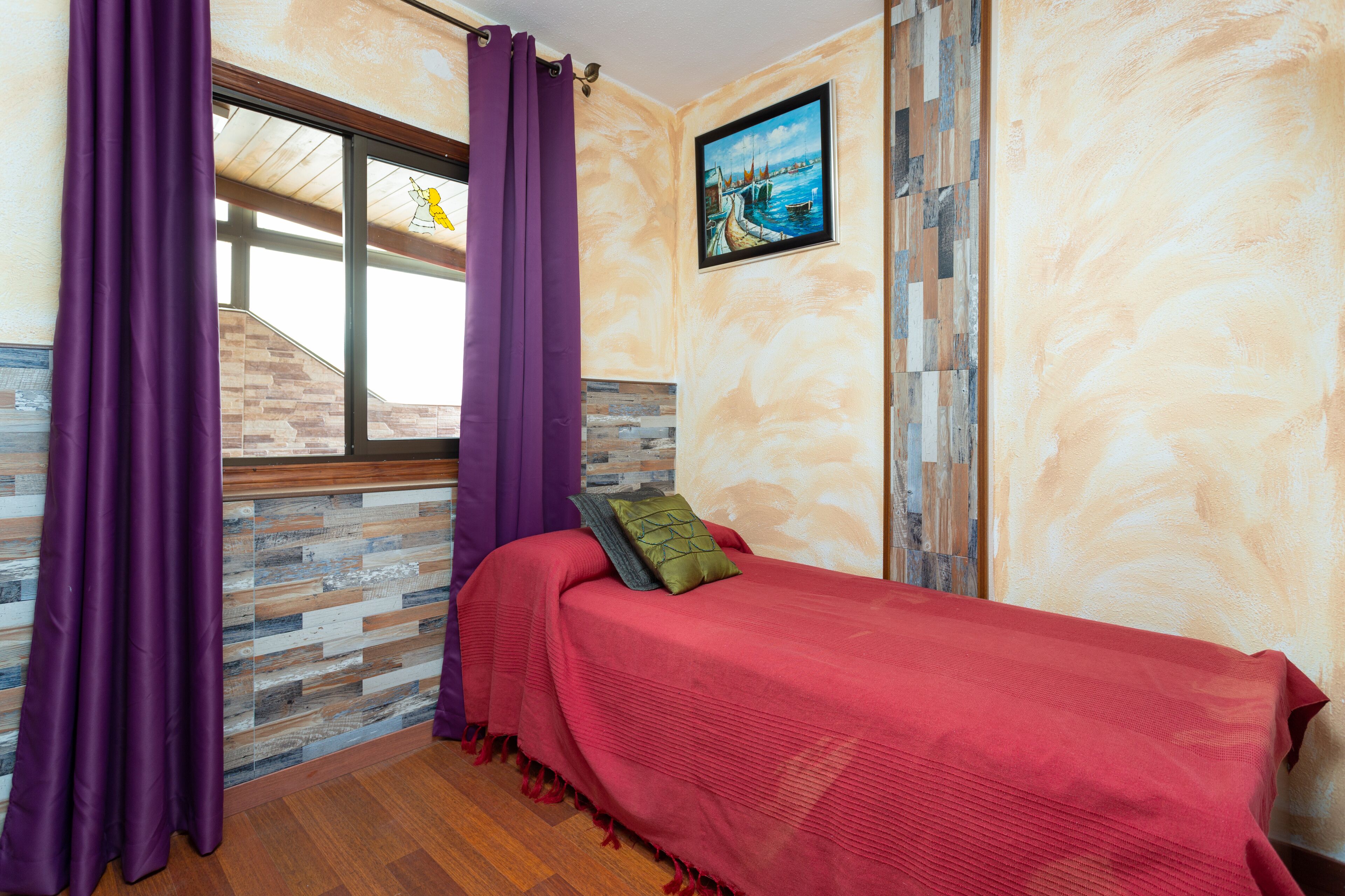 Ein Schlafzimmer mit Holzboden, einem Bett mit roter Bettwäsche, violetten Vorhängen und einem Gemälde, das eine Küstenlandschaft zeigt.