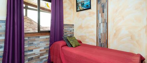 Ein Schlafzimmer mit Holzboden, einem Bett mit roter Bettwäsche, violetten Vorhängen und einem Gemälde, das eine Küstenlandschaft zeigt.