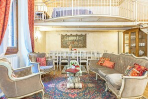 Una habitación amplia con techo alto, balcón, zona de comedor con sillas blancas y un cuadro grande en la pared.
