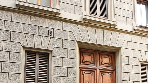 Eine Gebäudefassade mit einer großen Holztür, Steinmauern und einem Fenster mit Rollläden.