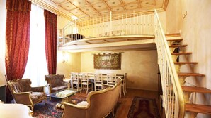 Ein Zimmer mit Esstisch, zwei Stühlen, einer Couch, einem Glastisch, einem gemusterten Teppich, einem Bild an der Wand und einer Treppe mit Geländer.