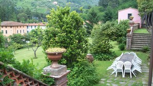 A lush garden with a large tree, a brick planter, and white plastic chairs. - Noble villa for 16 people, with park and pool in Tuscany  (Nocchi)