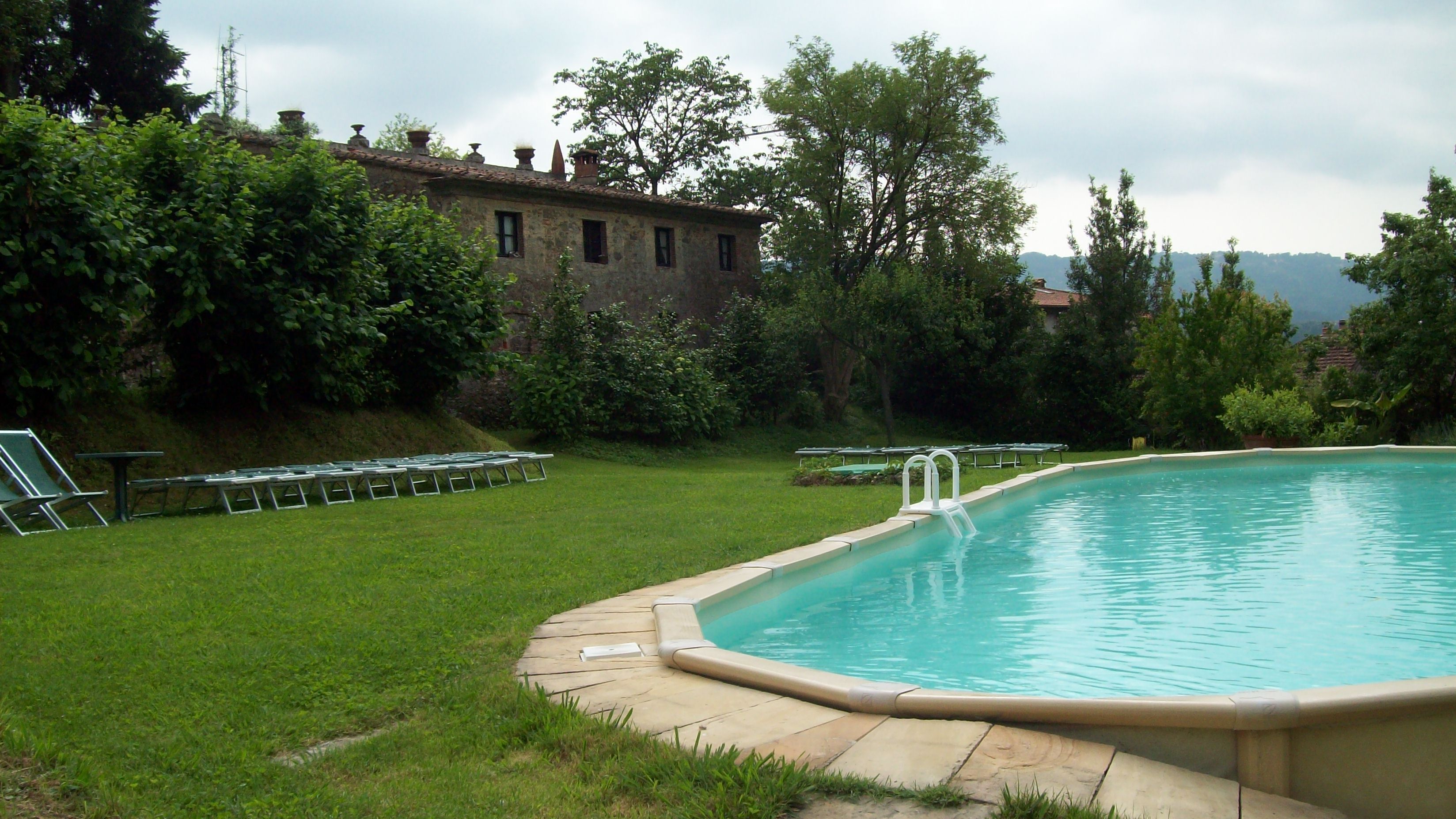 A swimming pool with a grassy area and lounge chairs, surrounded by trees and a building in the background.