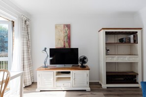 A room with a television on a wooden cabinet, a bookshelf, and a window with curtains.