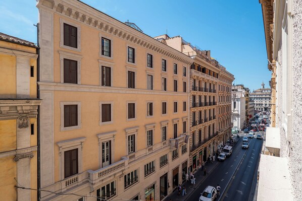 A multi-story building with a row of windows and balconies, a street with cars and pedestrians, and a clear sky.