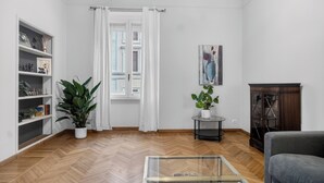 A living room with a herringbone floor, a glass coffee table, a dark armchair, a bookshelf, and a potted plant.
