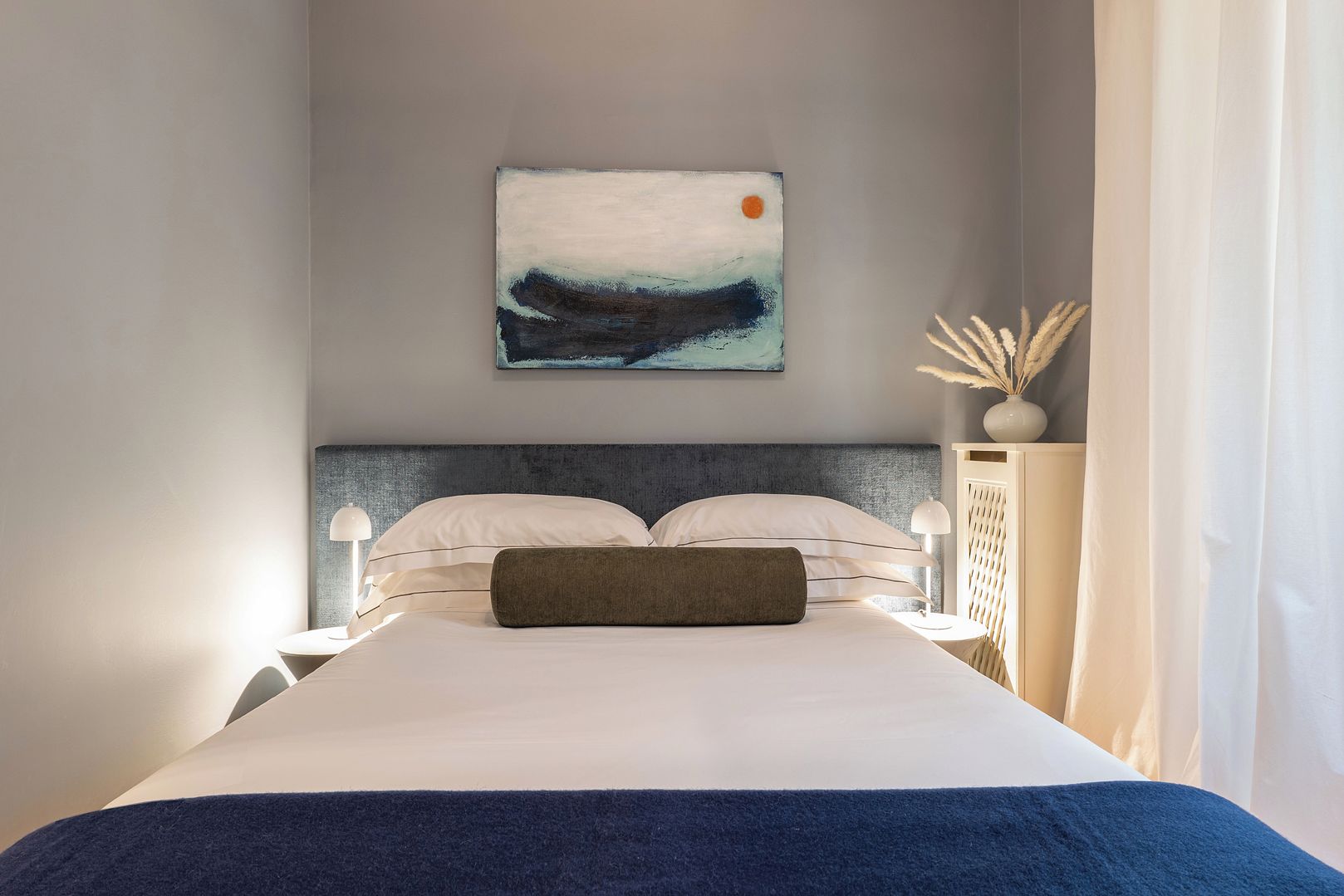 Habitación de hotel con cama, mesita de noche, lámpara, un cuadro y un jarrón con plantas secas.