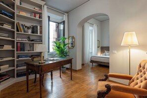 A room with a wooden desk, bookshelves, a potted plant, a sofa, and a lamp.